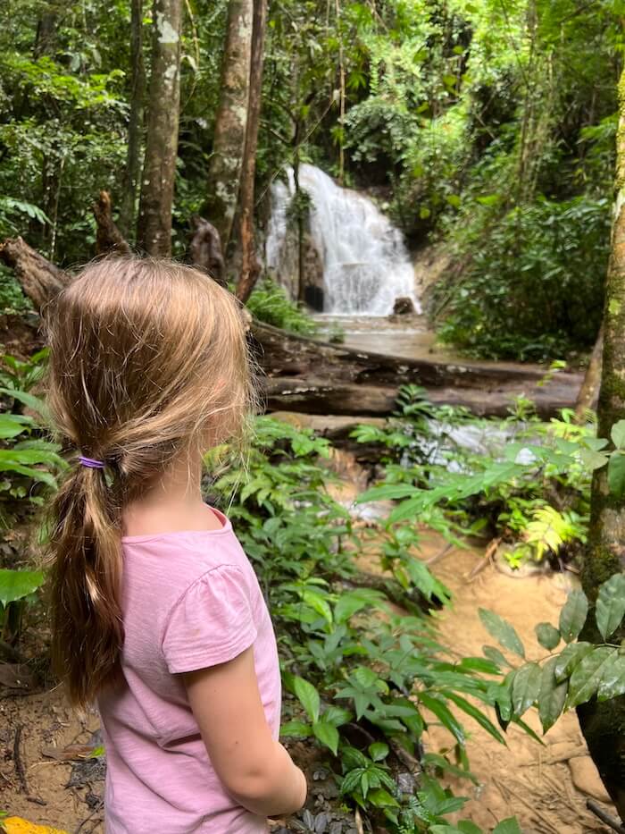 A young girl with a ponytail looks at a small cascade waterfall and stream in the lush, remote jungle of Doi Luang National Park.