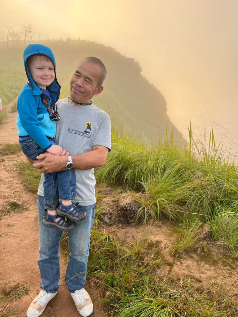 A kind, older Thai man (the Ukrist Farm owner) smiling while carrying a young child on his hip down the muddy, grassy trail at Phu Chi Fa after the sunrise hike.