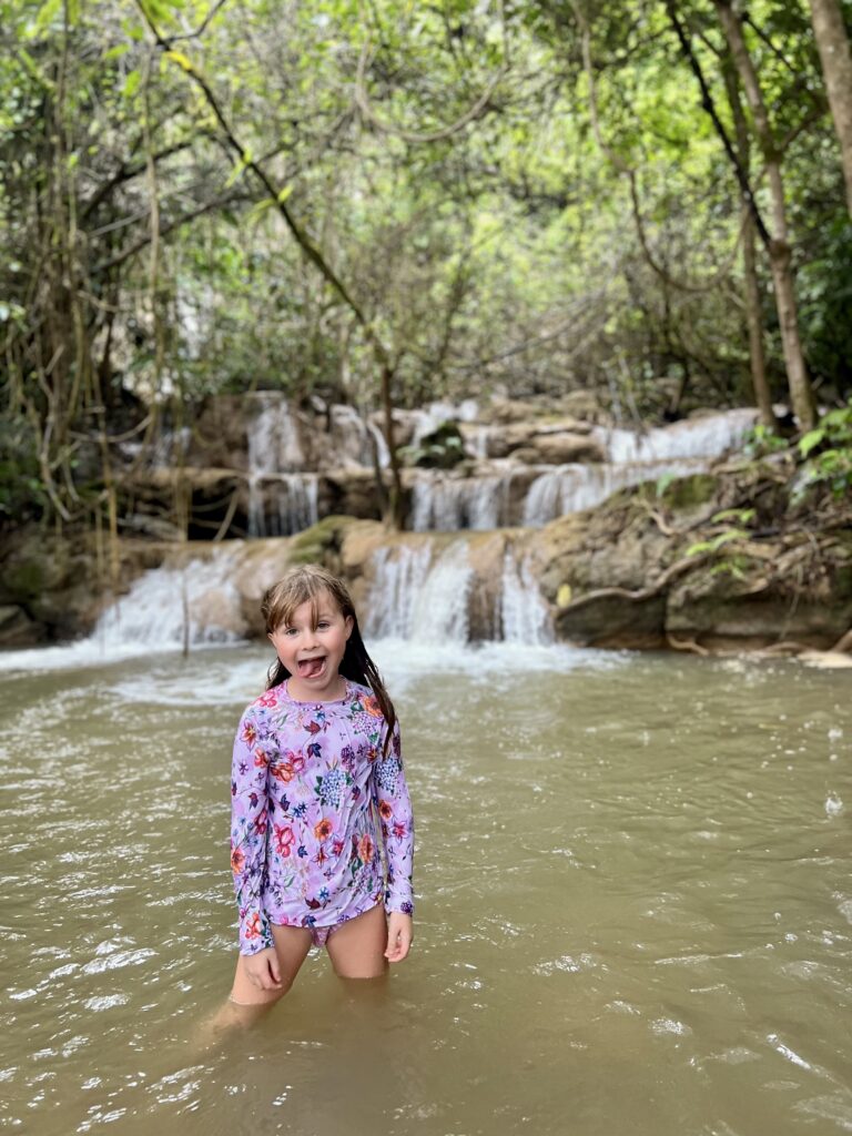 Young girl in a rash guard swimming and playing in the shallow pool at the base of a tiered waterfall in the Umphang jungle.