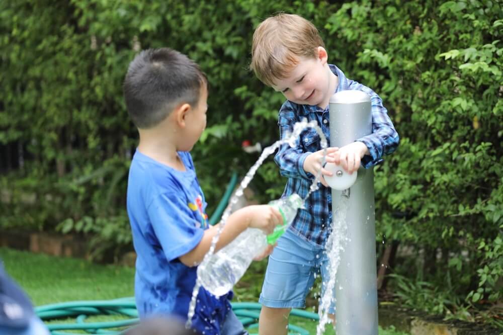 Two young boys playing happily with water at an outdoor fountain in a playground, illustrating hands-on learning and social play during worldschooling.