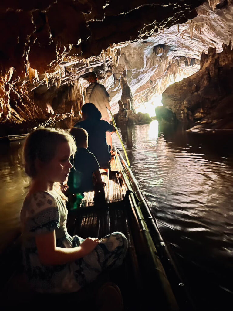 A young girl sitting on a bamboo raft inside a massive limestone cave with stalactites, heading toward a bright opening of light.
