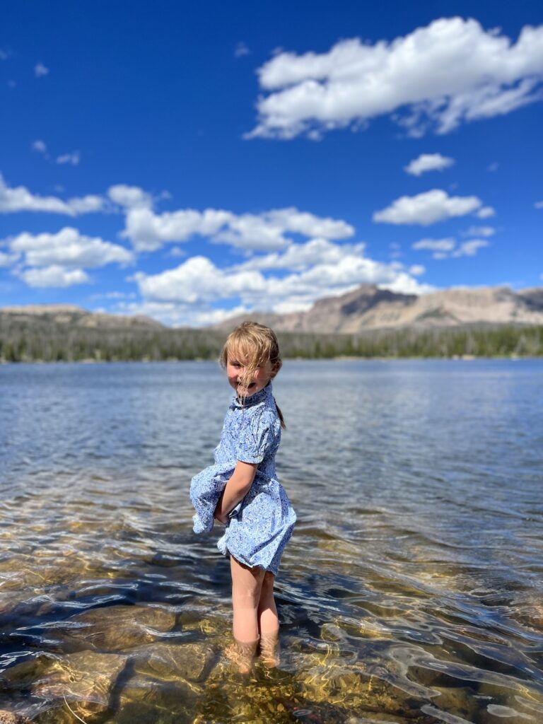 Young girl in a blue dress wading in a clear, shallow alpine lake with a background of mountains and a bright blue sky.
