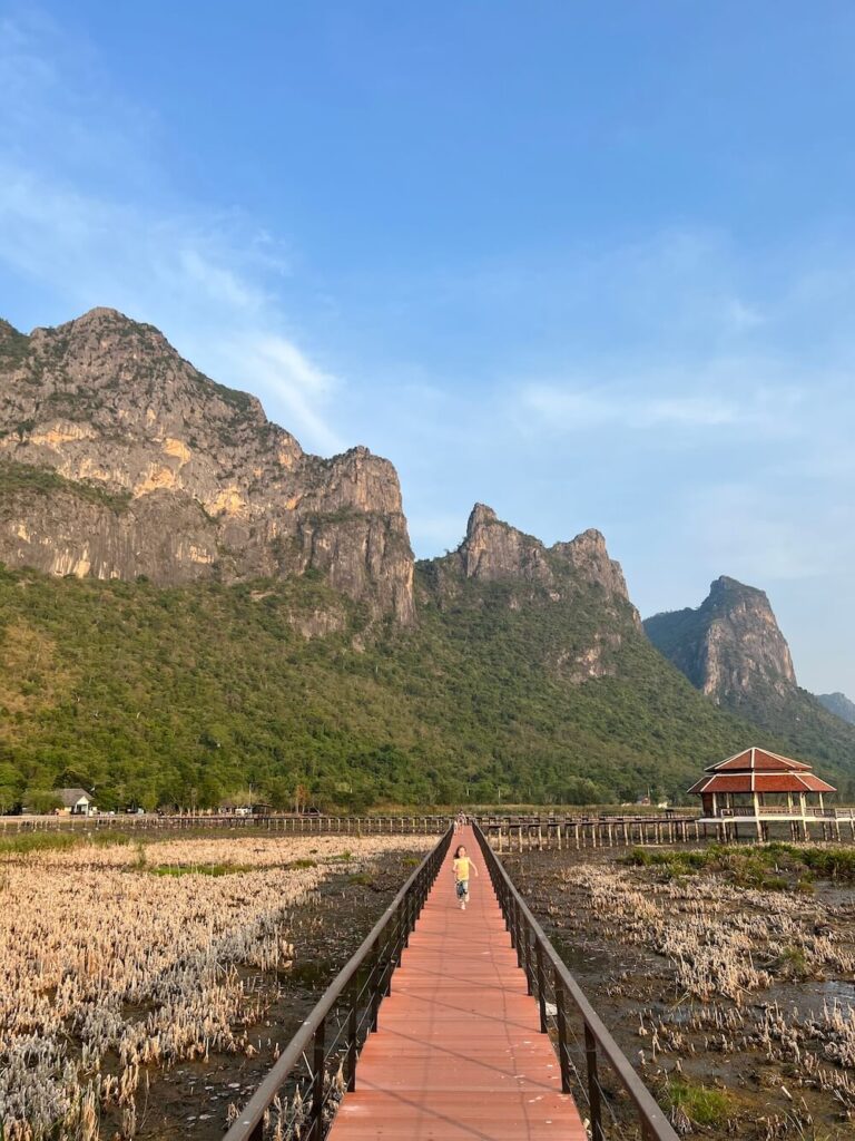 A child walking along the red wooden Bueng Bua Boardwalk, stretching across the dried marshland towards a pavilion, backed by the sheer, jagged limestone karst mountains of Khao Sam Roi Yot National Park.