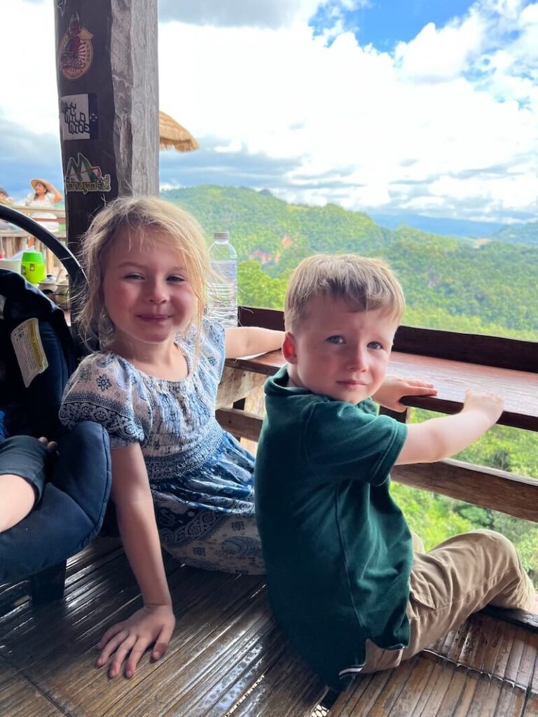 A young boy and girl sitting on a bamboo floor at a ridgetop restaurant in Ban Jabo, with a lush mountain valley in the background.