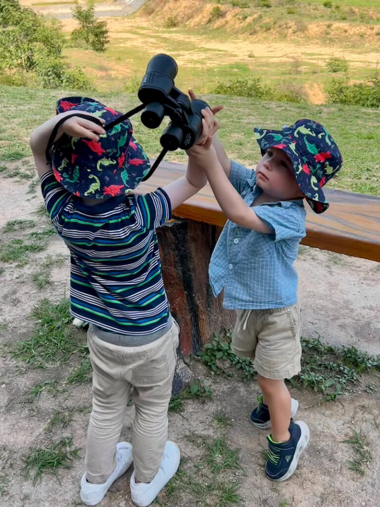 Two young children wearing sun hats and collaborating to hold a pair of large binoculars, intently searching the horizon for wildlife.