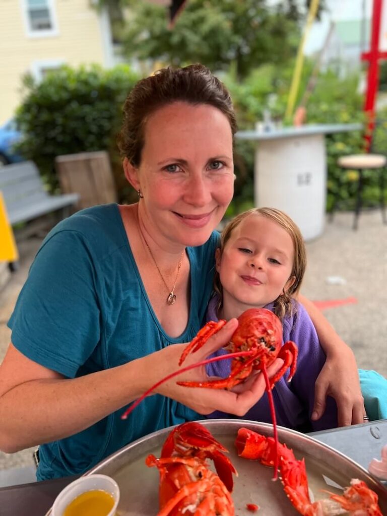 HOME Mother and young daughter smiling while holding a whole cooked red lobster outdoors.