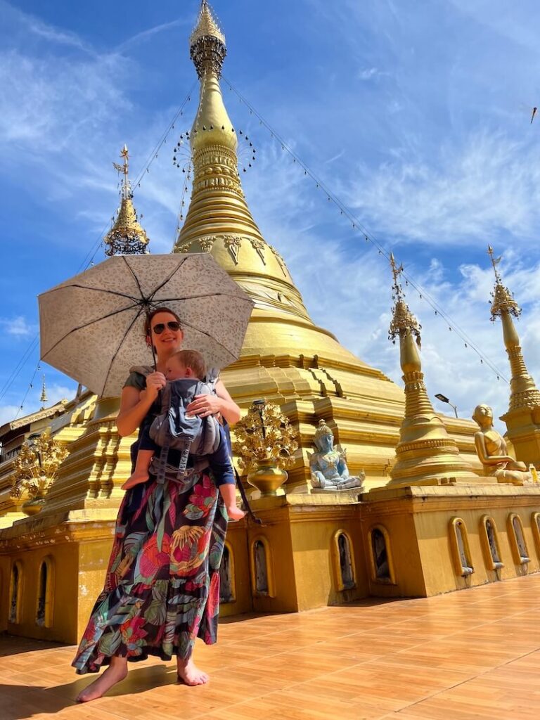 Woman in a flowered dress and baby carrier holding an umbrella in front of a large golden pagoda under a blue sky.