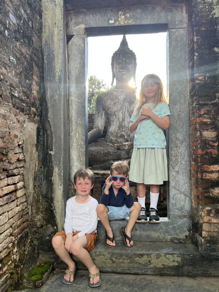 Three young children sitting in the doorway of a Sukhothai temple ruin with a large Buddha statue visible behind them, emphasizing family travel.