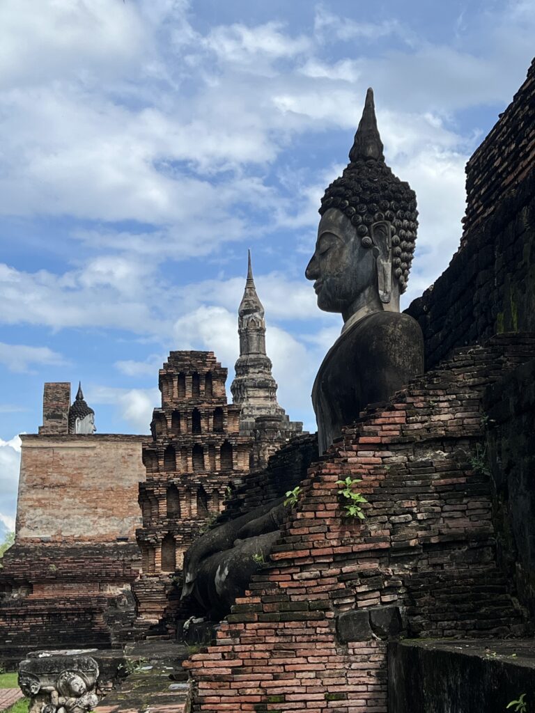 Profile view of a large, seated Buddha statue resting against brick ruins and layered spires in the Central Zone of Sukhothai Historical Park.