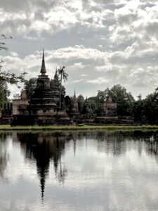 Ancient temple spires and ruins of Wat Mahathat reflecting clearly in the still water of the surrounding reservoir in Sukhothai Historical Park.