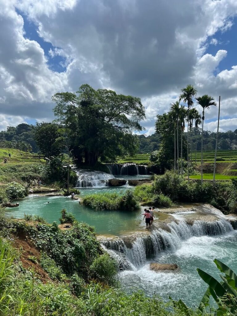 Multi-tiered, cascading waterfalls with bright turquoise pools over rock terraces, surrounded by lush green rice paddies and tropical jungle in Sumba, Indonesia.