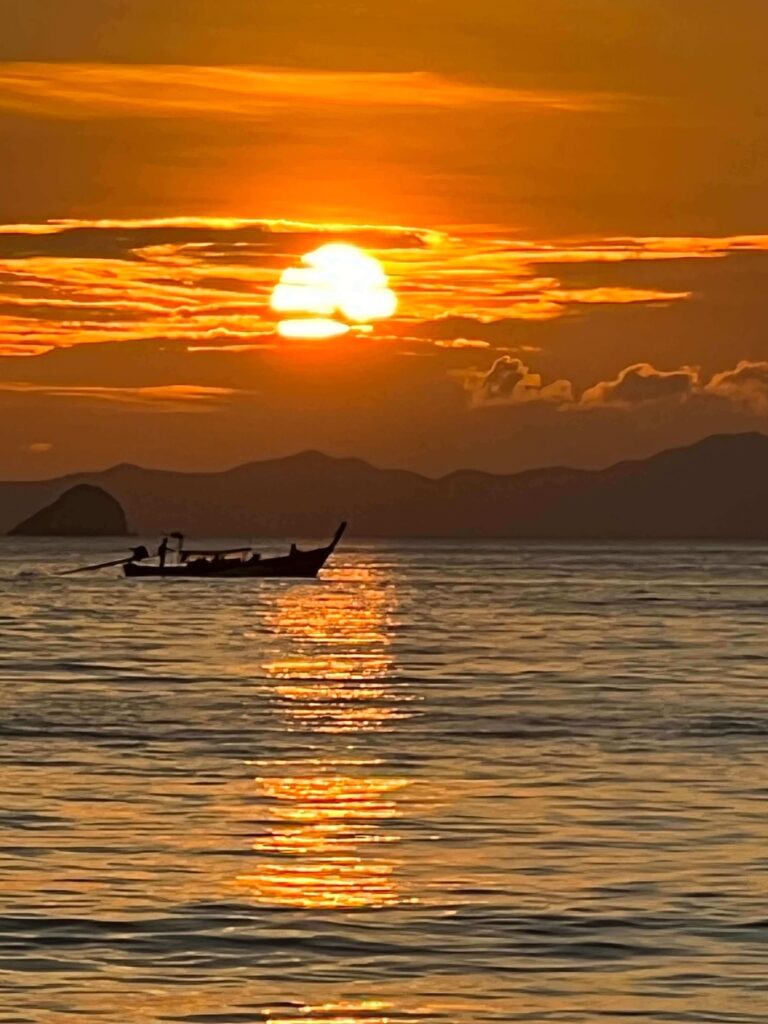 Thai longtail boat silhouetted against a brilliant orange sunset over the Andaman Sea with coastal mountains in the background.