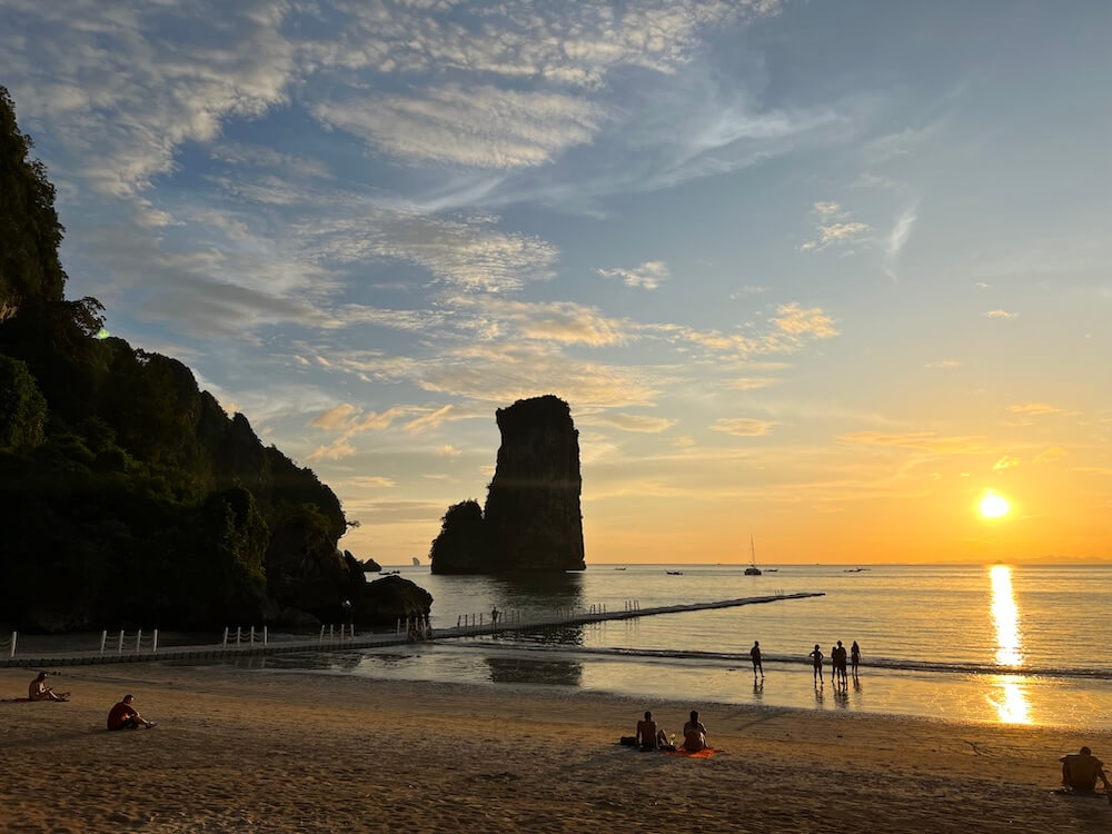 HOME A breathtaking tropical sunset over a sandy beach in Thailand, featuring a striking karst island formation in the distance and silhouettes of people enjoying the golden hour.