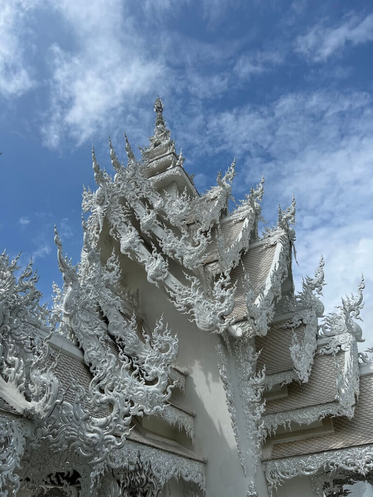 A detailed close-up of the intricately carved white exterior of the Wat Rong Khun (White Temple) in Chiang Rai, Thailand, set against a bright blue sky.