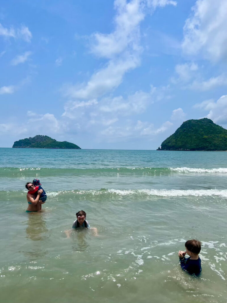 A family swimming in the warm, shallow, calm turquoise water of Ao Manao beach, with two steep, forested limestone islands visible in the background.