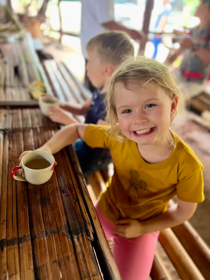 A young girl smiling while holding a cup of tea at a rustic bamboo counter in the Ban Mae Klang Luang coffee roasters on Doi Inthanon.