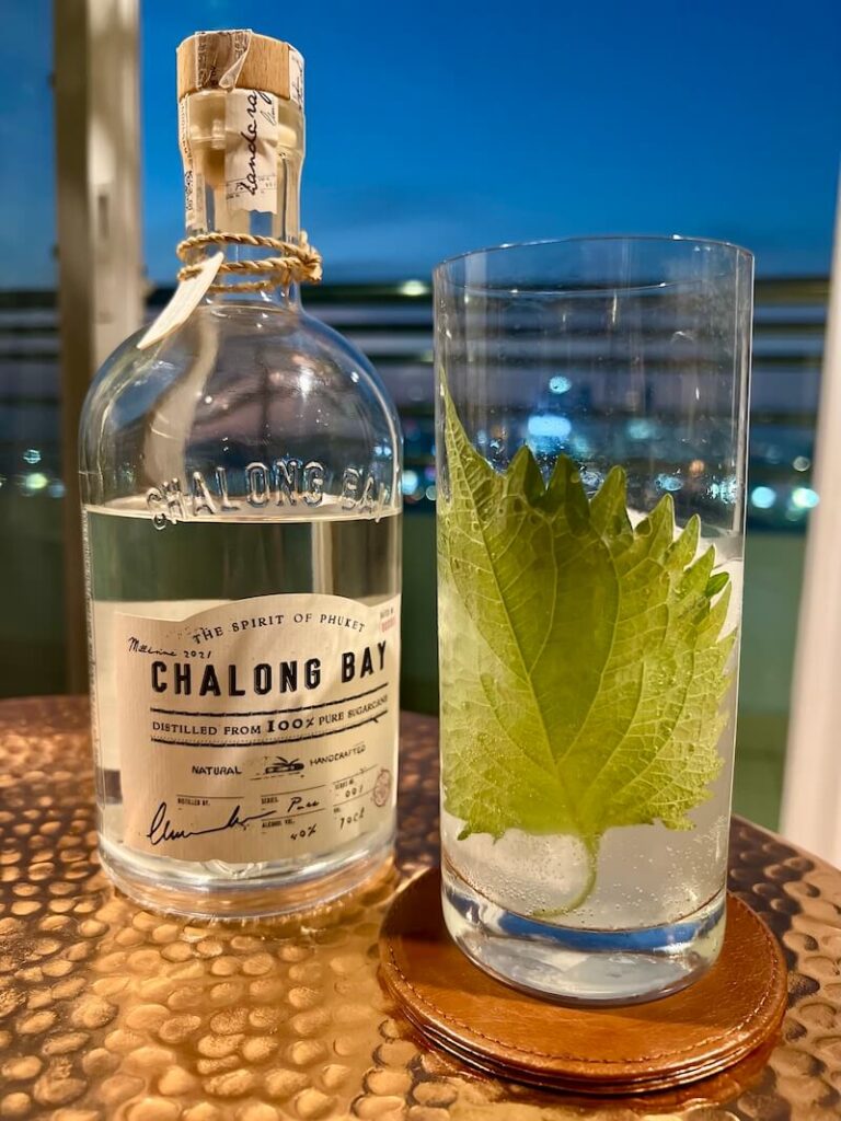 A highball cocktail featuring a large green shiso leaf pressed against the glass, standing next to a bottle of Chalong Bay Thai Sugarcane Rum on a copper table overlooking the Bangkok skyline at dusk.