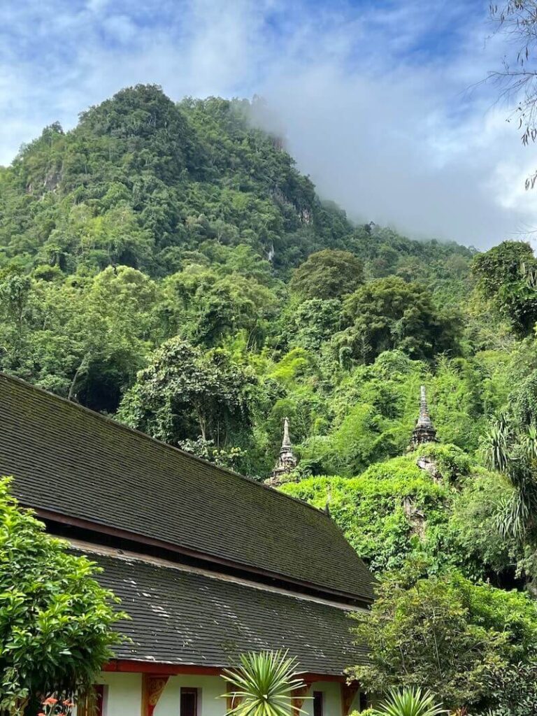 Alt Text: A view of the weathered, moss-covered chedis and temple roof at the entrance of the Chiang Dao Temple Caves, with mist-shrouded limestone cliffs in the background.