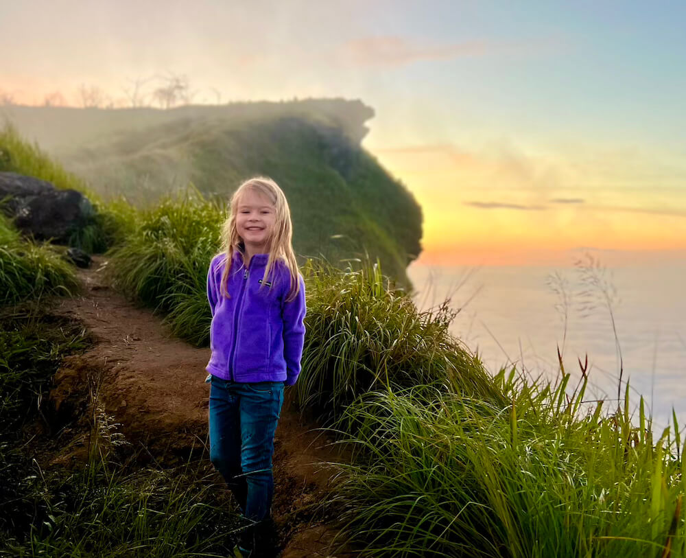 Phu Chi Fa Trail Girl Sunrise Sea of Fog Young girl smiling on the dirt trail at Phu Chi Fa summit, with the sun rising over the dense sea of fog and grass-covered cliffs behind her.