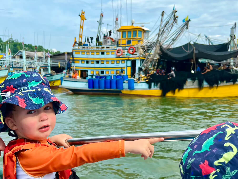 Toddler in a life vest pointing toward large, colorful wooden fishing boats docked in the Chumphon River, Thailand.