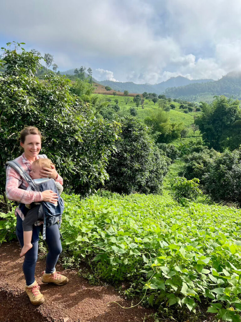 A woman hiker carrying a sleeping baby in a front-facing carrier stands on a dirt path overlooking lush green hills and mountains in Chiang Dao, Thailand.