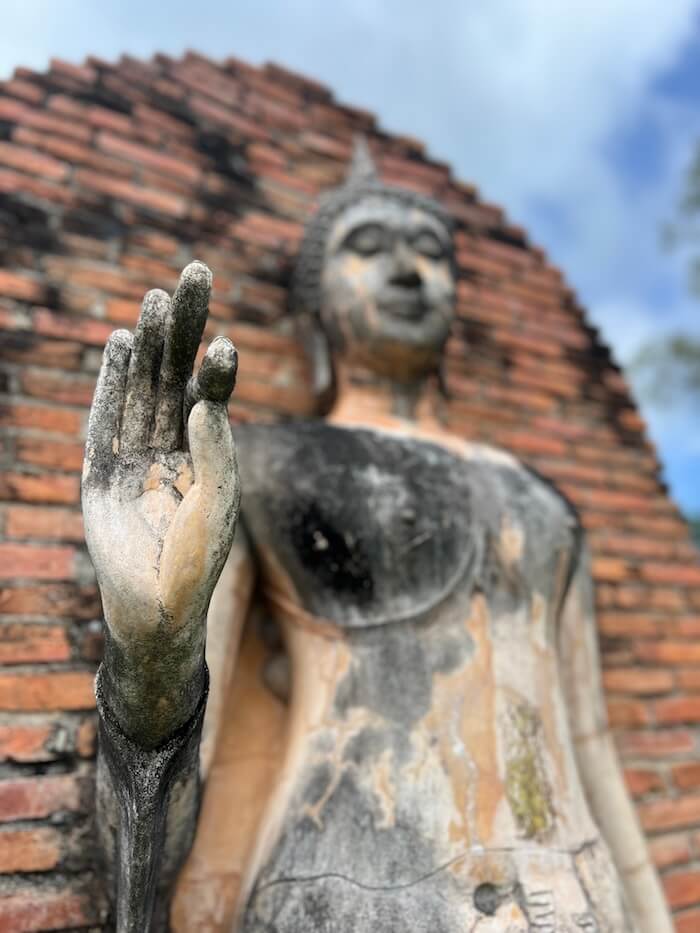 Standing Buddha statue with hand raised in peace gesture against brick ruins