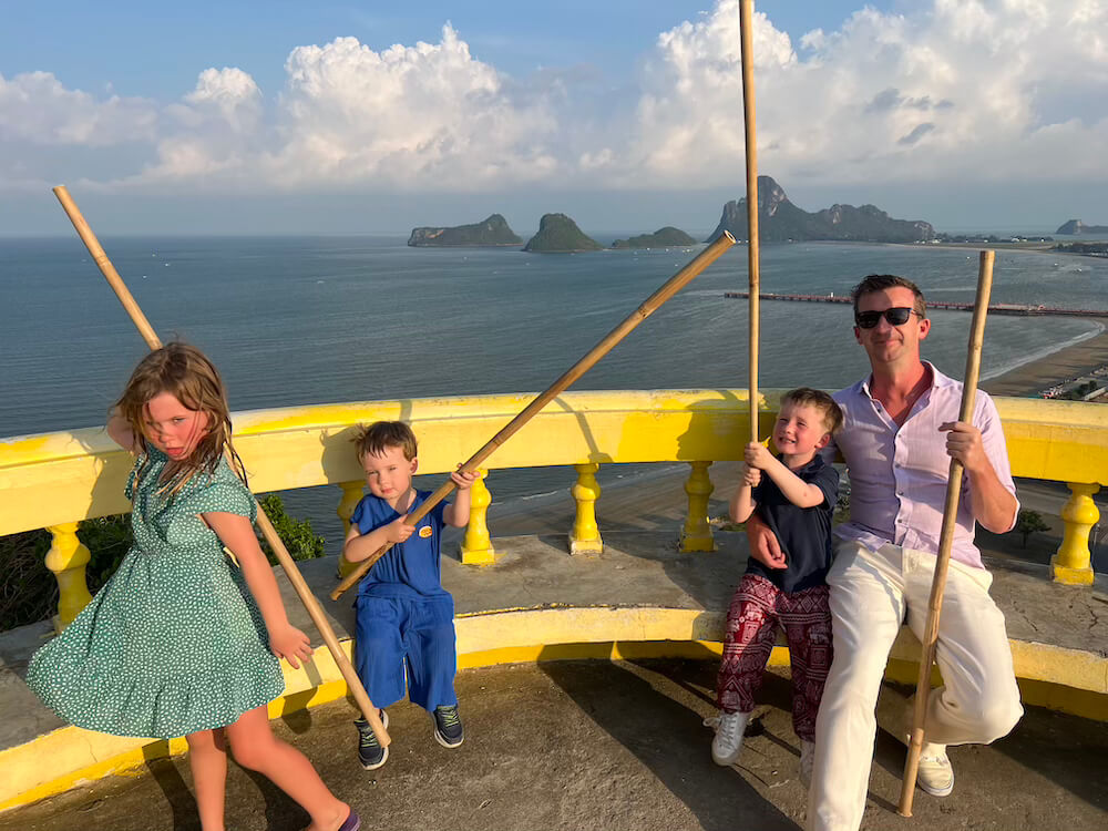 A family standing at the summit of Wat Khao Chong Krachok overlooking Ao Prachuap, each holding a long wooden stick for defense against the macaque monkeys.