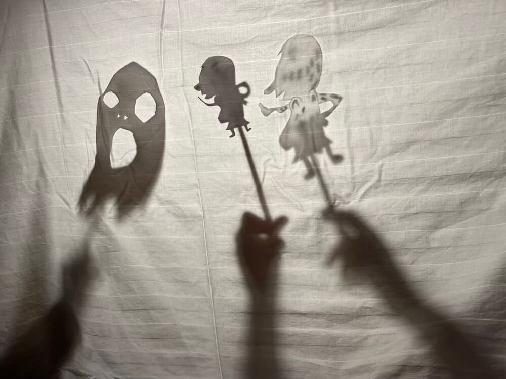Homemade shadow puppets of a ghost and two children characters being performed behind a white sheet by children's hands.