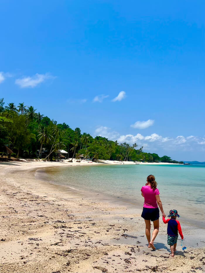 A mother and young child walking hand-in-hand along the curved, white sand beach of Koh Talu Island, Thailand, with clear, shallow water and lush tropical jungle in the background.