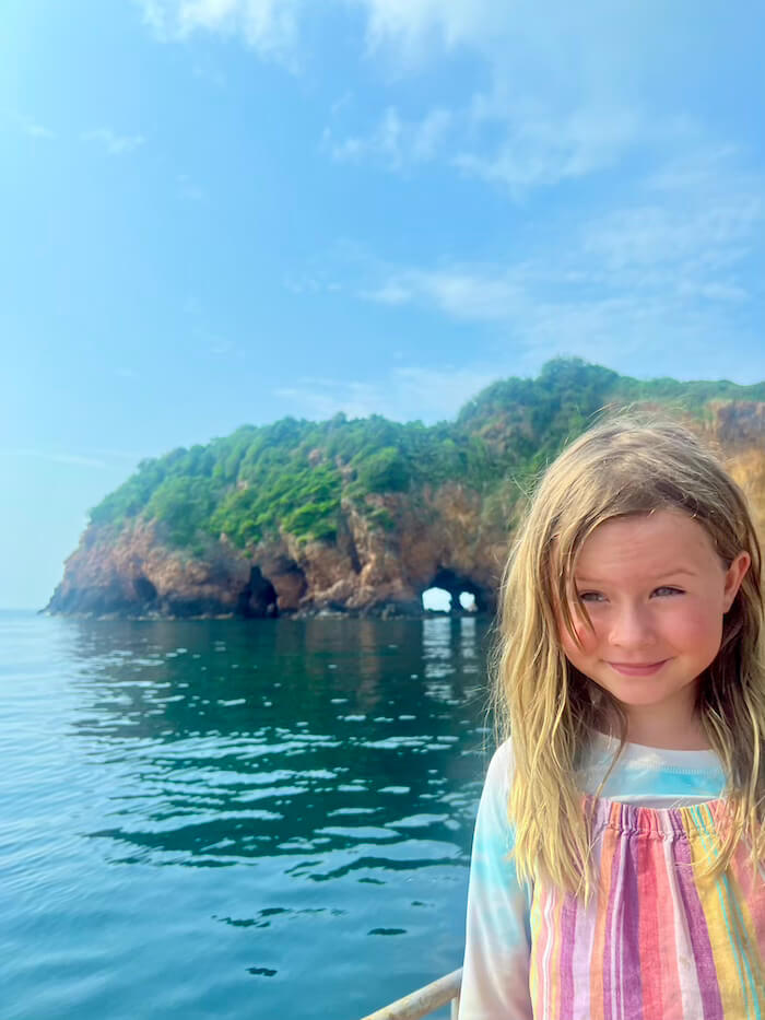 A young girl smiling on a boat with the famous hole (archway) in the rocky, green cliff face of Koh Talu Island visible over the water in the background.