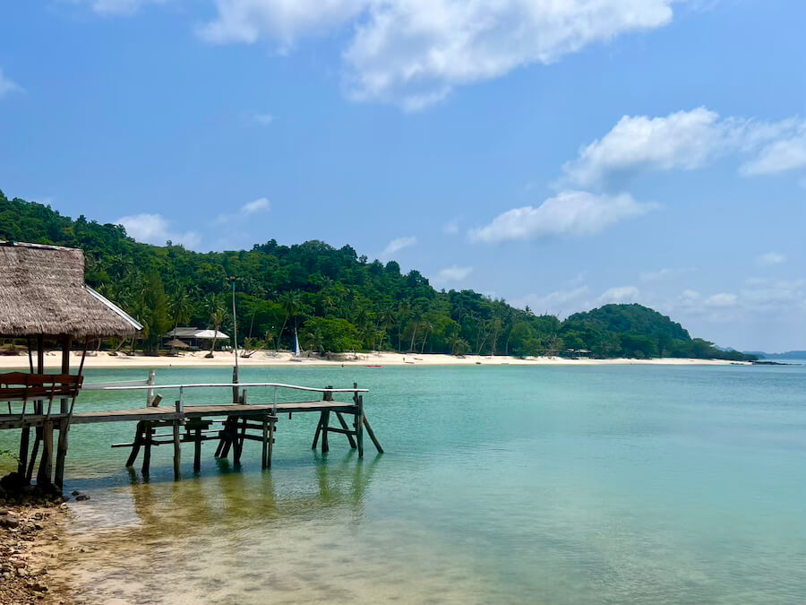 A wooden pier with a small thatched hut extends into a calm, shallow turquoise bay on Koh Talu Island, with a white sand beach and dense green jungle shoreline visible in the distance.