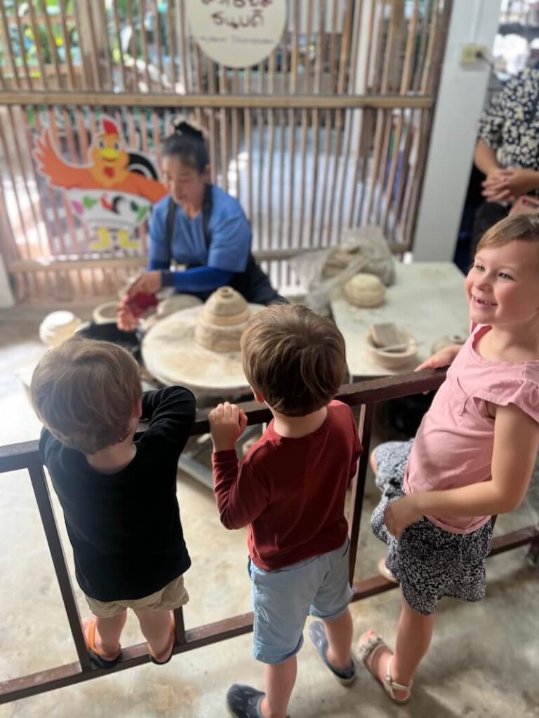 Three young children lean against a wooden railing, watching a local artisan demonstrate how to use a pottery wheel in an open-air workshop.