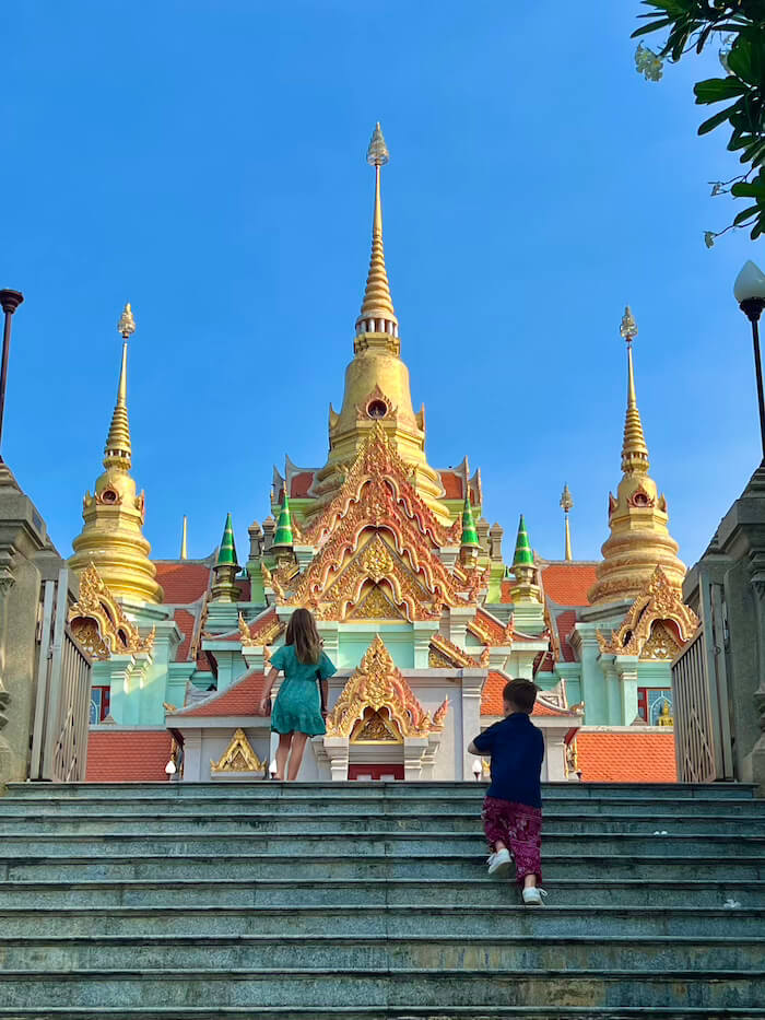 Two children climbing the wide stone steps toward the elaborate, multi-spired golden pagoda of Phra Mahathat Chedi in Bang Saphan, Thailand.