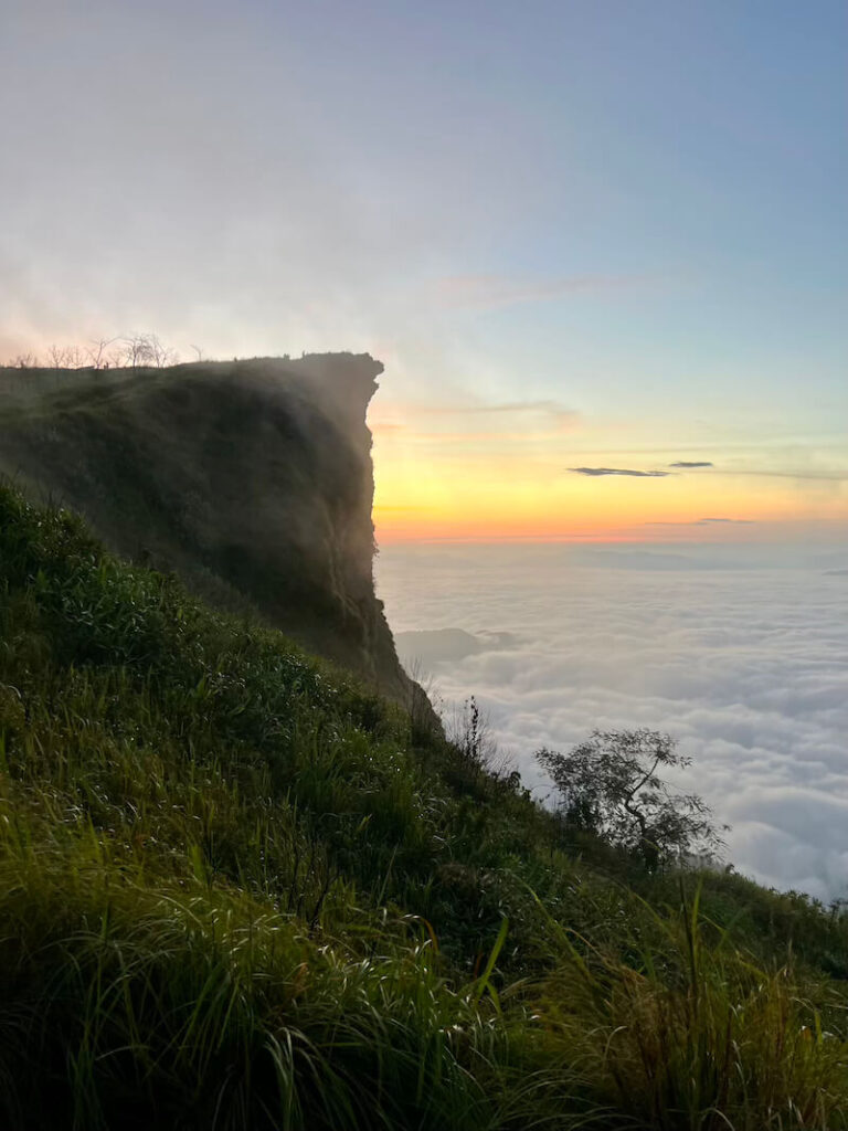 Dramatic view of the sheer cliff face at Phu Chi Fa summit overlooking a dense, white sea of clouds with the orange sunrise horizon in the distance.
