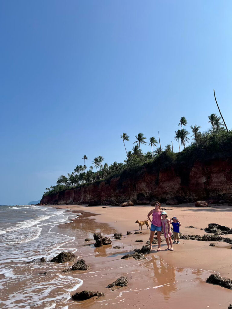 A mother and two children stand on the red sand beach beneath the towering, dark red laterite cliffs of Pha Daeng, Bang Saphan Noi, Thailand, with a friendly stray dog nearby.