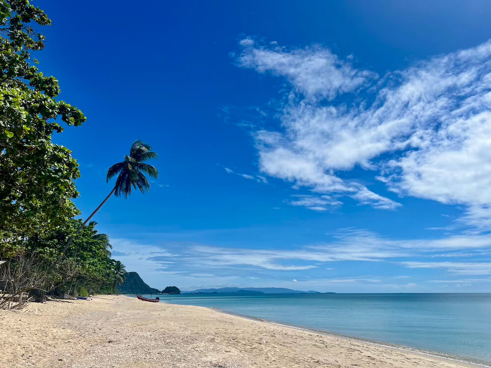 View of a secluded, uncrowded beach with a single palm tree, clear water, and distant mountains on the coast of Khanom, Thailand.