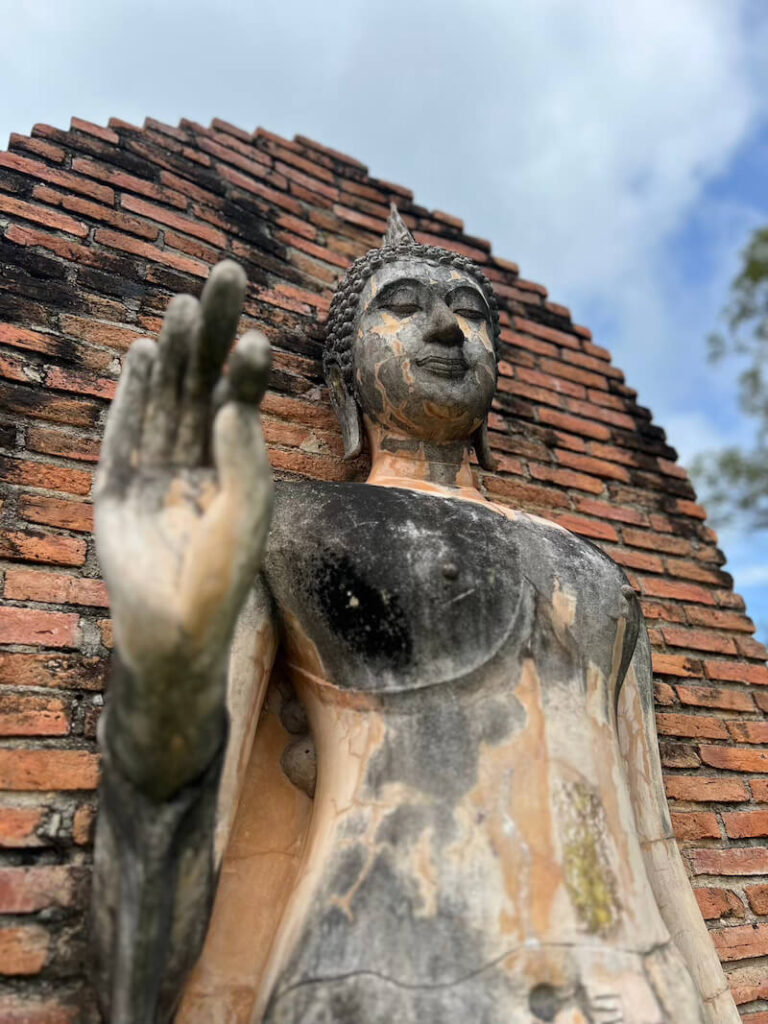 A weathered, standing Buddha statue with its hand raised in the Abhaya Mudra (do not fear) pose, set against an ancient brick ruin at Sukhothai Historical Park.