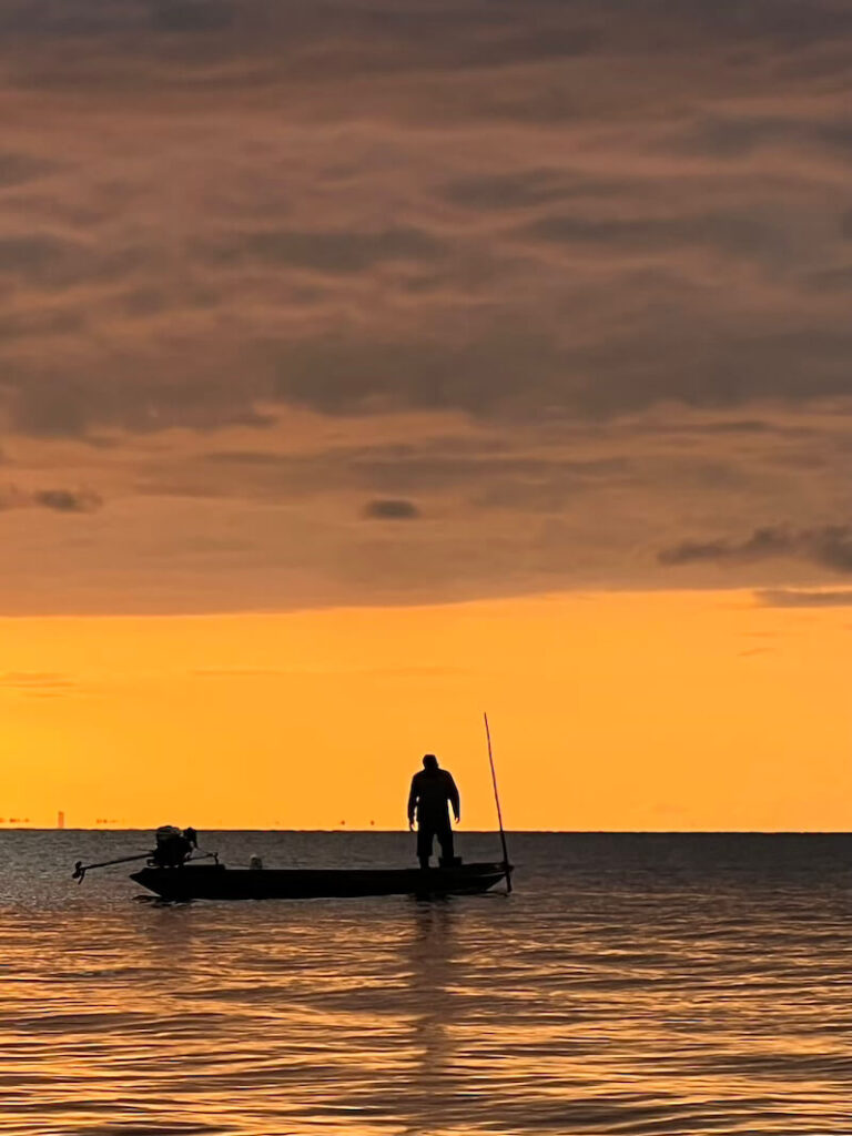 Silhouette of a fisherman standing in a small boat against a bright orange and red sunset on Songkhla Lake near Thale Noi.