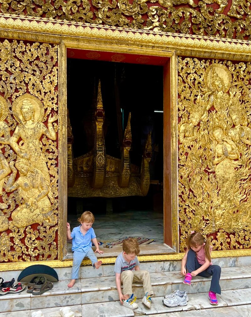 Three young children sitting on the marble steps of a golden, intricately carved Thai temple entrance, putting on their shoes.