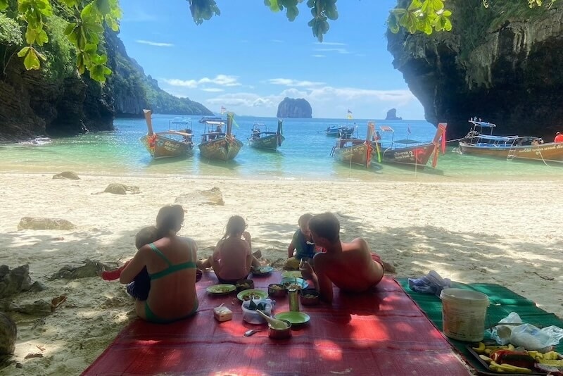A family having a picnic on a beach in Thailand with longtail boats in the turquoise water.