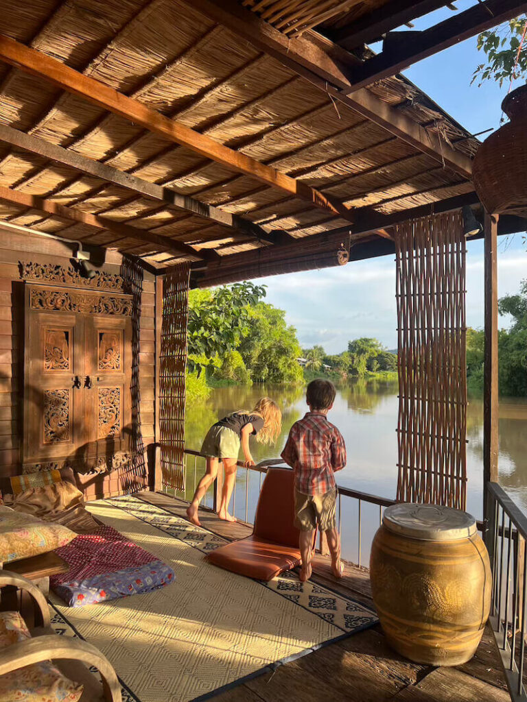 Two children look over the railing of a covered wooden deck built over a calm river in Thailand, with lush jungle foliage in the background.