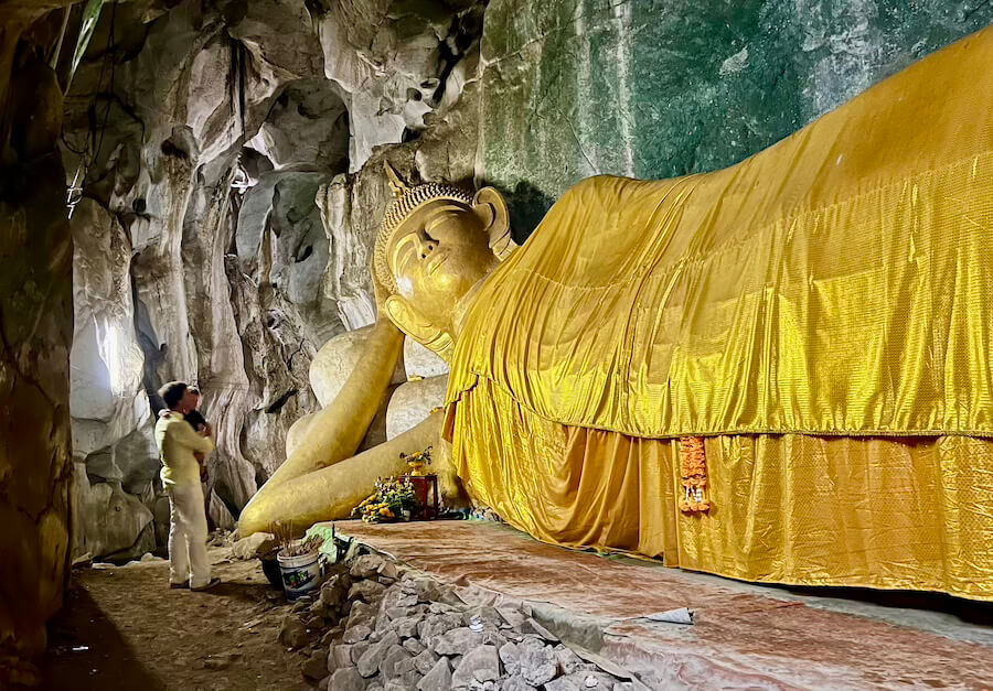 A large, gilded Reclining Buddha statue inside the Tham Phra Non limestone cave at Wat Ao Noi. The statue is hundreds of years old, and a visitor stands nearby, showing the immense scale.