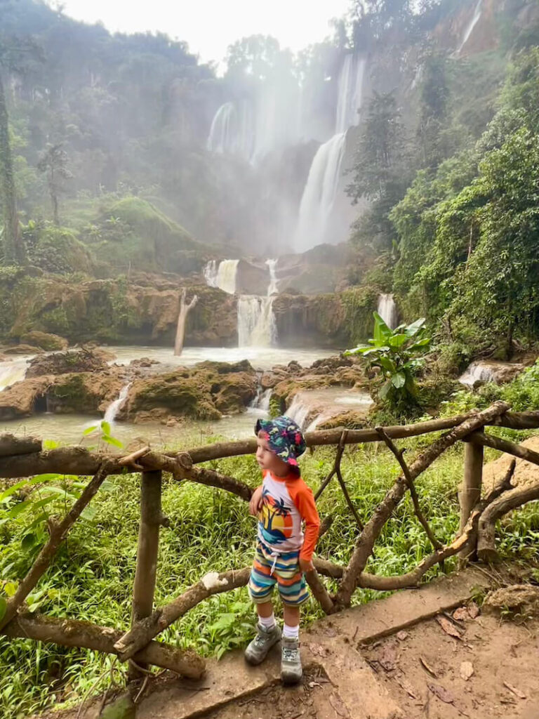 Young child standing on a viewing platform overlooking the massive, multi-tiered cascade of Thi Lor Su Waterfall in Umphang, Thailand.