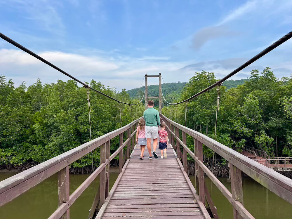 Father and two children walking away on a long wooden suspension bridge over a canal in the Thung Ka Bay Mangrove Forest, Chumphon, Thailand.