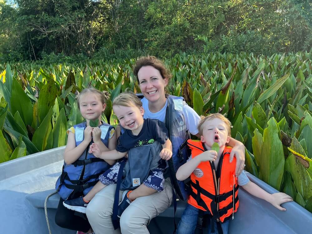 A mother and three young children in a boat, wearing life vests, exploring a lush green jungle waterway as part of the Touch Grass Tour family travel blog