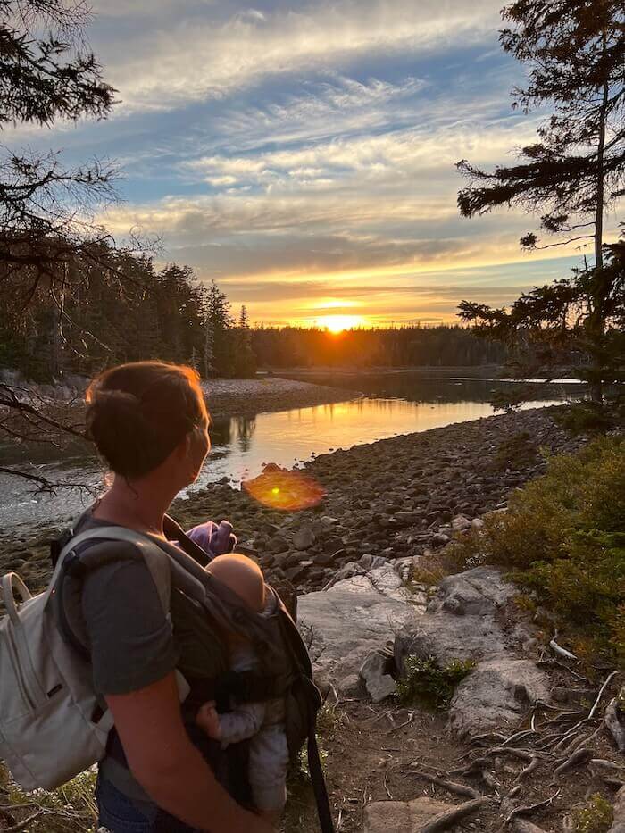 Mother hiking with baby in carrier watching sunset over water and rocky shore