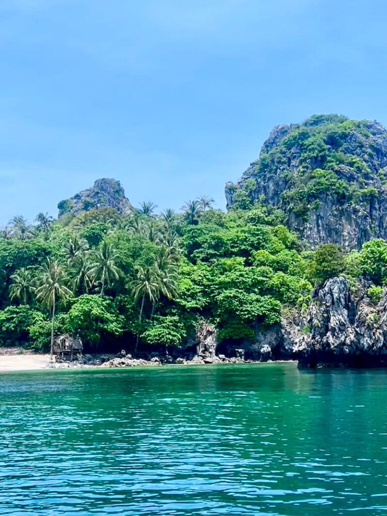 Rocky limestone island covered in bright green tropical jungle and palm trees rising from clear turquoise water in the Gulf of Thailand.