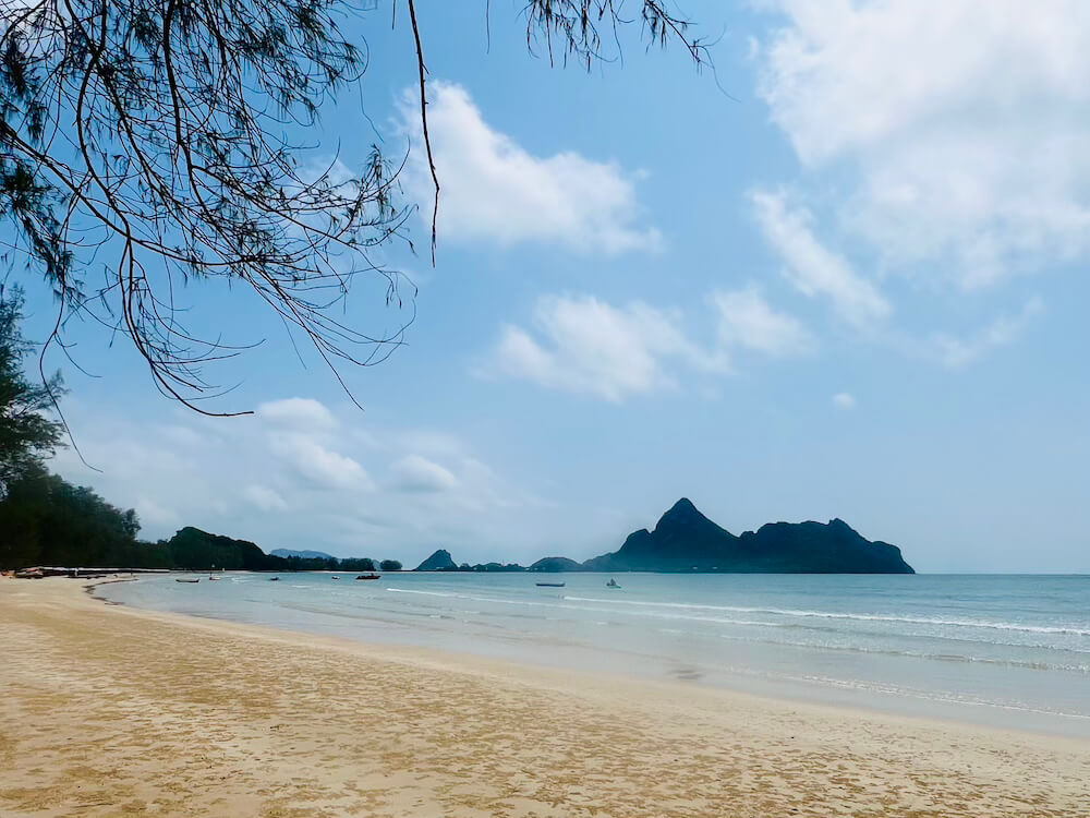A wide, empty expanse of sandy beach on the Gulf of Thailand, showing forested limestone karsts in the distance and very few boats, illustrating the unspoiled Thai Riviera.