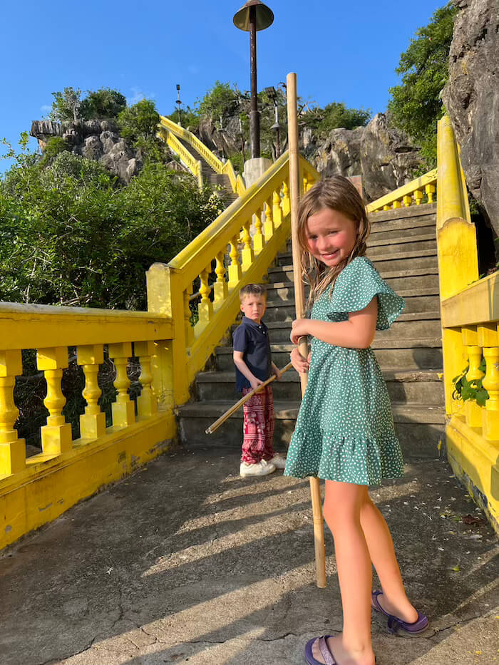 Two children standing at the base of the steep concrete staircase leading up to Wat Khao Chong Krachok, distinguished by bright yellow railings, both holding wooden macaque defense sticks.