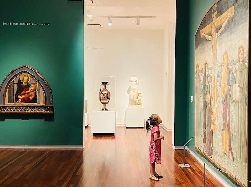Alt Text: A young girl in a pink dress standing in an art gallery, looking up at a large classical painting of a religious scene surrounded by green walls and artifacts.